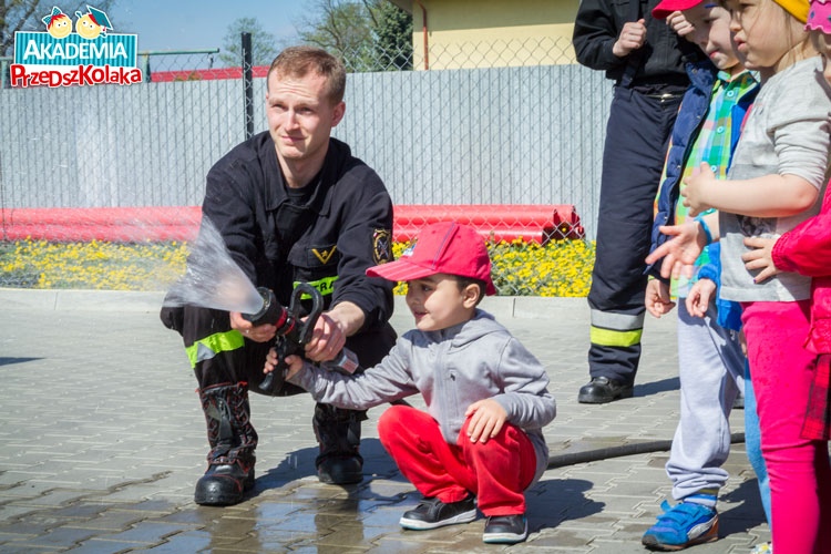 Przedszkolak z pomocą strażaka leje wodą z węża strażackiego.