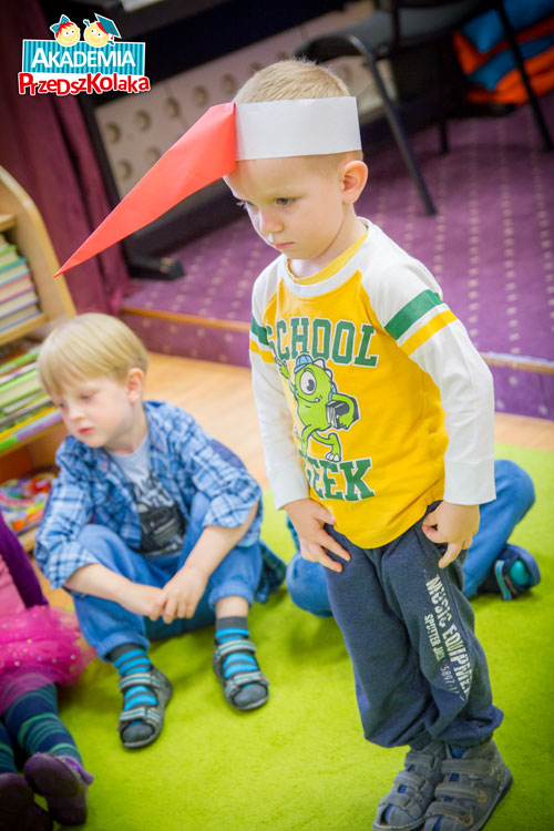Przedszkolak w zabawie z papierowym, czerwonym dziobem na głowie udaje bociana.