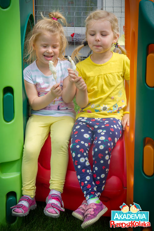 Dwie uśmiechnięte dziewczynki siedzą na zabawce i jedzą watę cukrową. 