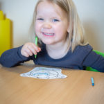 Dziewczynka się uśmiecha. Siedzi przy stole i koloruje rysunek ziemi.