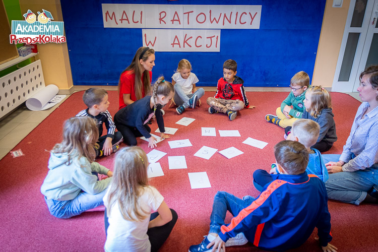 Grupa przedszkolaków siedzi na dywanie przed rozrzuconymi kartkami formatu A4. Na kartkach napisane są pojedyncze litery.
