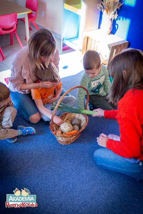 Przedszkolaki z nauczycielką siedzą na dywanie i przyglądają się zawartości koszyka. W środku leży dużo różnych warzyw. Seler, marchew, buraki, por.