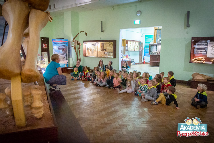 Widok od strony podestu na którym stoi wielki szkielet dinozaura i siedzi przewodniczka. Widać całą grupę dzieci siedząca na podłodze i słuchająca pracownicy muzeum.