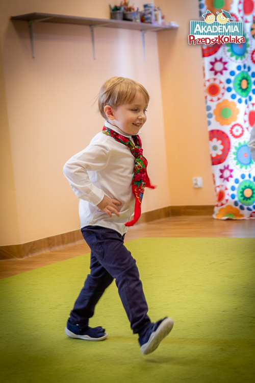Przedszkolak dziarsko kroczący przez salę. Chłopiec wypełnia cały kadr. Biała koszula i czerwona chusta na szyi. Uśmiechnięty.