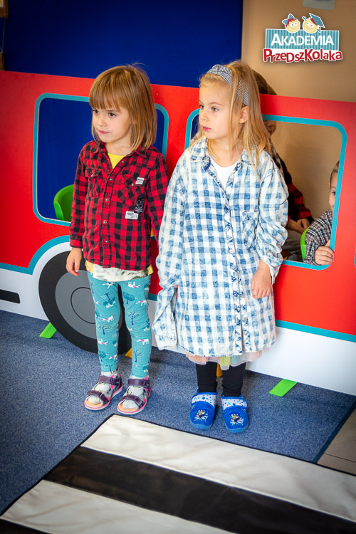 Dwoje przedszkolaków, dziewczynki. Trzymając się za rączki stoją przed pasami przejścia ułożonych w sali przedszkolnej do ćwiczenia zasad obowiązujących pieszych w mieście. 