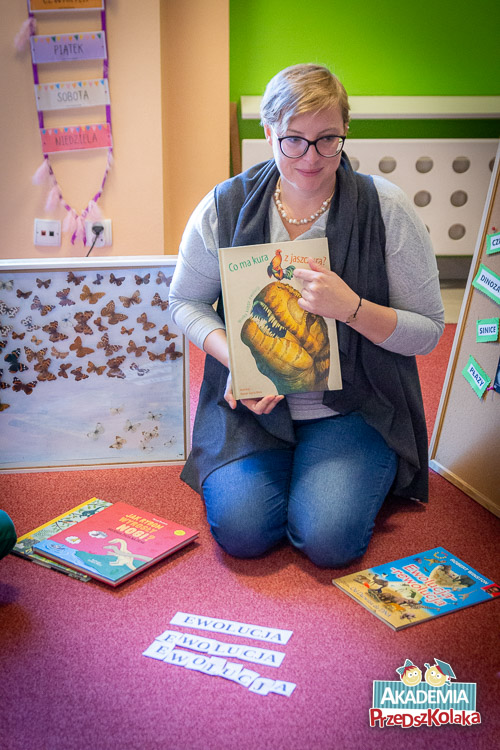 Nauczycielka znajduje się na dywanie, klęczy. Trzyma w rękach książkę Co ma kura z jaszczura. Po bokach, z lewej i prawej strony leżą inne książki. Na wprost leżą wyrazy EWOLUCJA, układane wcześniej przez przedszkolaków.