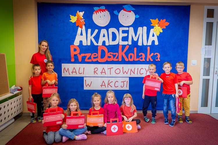 Zdjęcie grupowe. Cała grupa przedszkolna z przedszkola na Tarchominie. Wszyscy dumnie trzymają swoje teczki prac. Dzieci są ubrane w czerwone koszulki. Z dziećmi stoi nauczycielka. W czerwonej koszulce. 