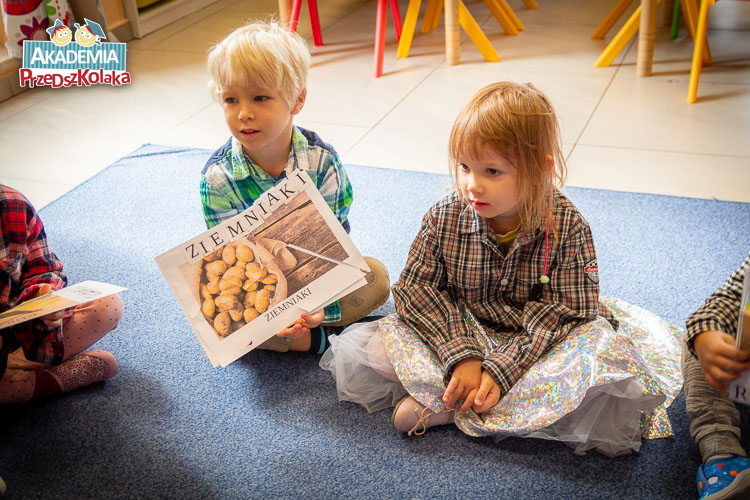 Przedszkolaki siedzące na dywanie trzymają kolejny kolaż z puzzli, tym razem przedstawiający ziemniaki. Obok przyklejony prawidłowy napis odpowiadający ilustracji. 