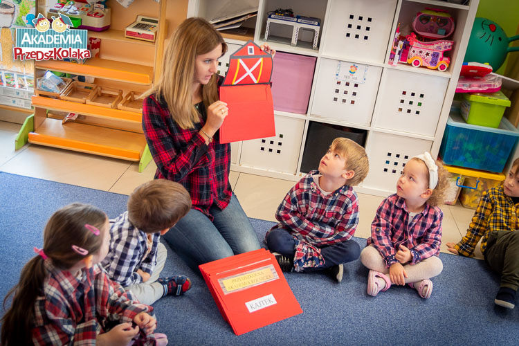 Pani nauczycielka z przedszkola na Białołęce pokazuje dzieciom siedzącym na dywanie, czerwoną teczkę która będzie służyła do zbierania materiałów związanych z projektem. 