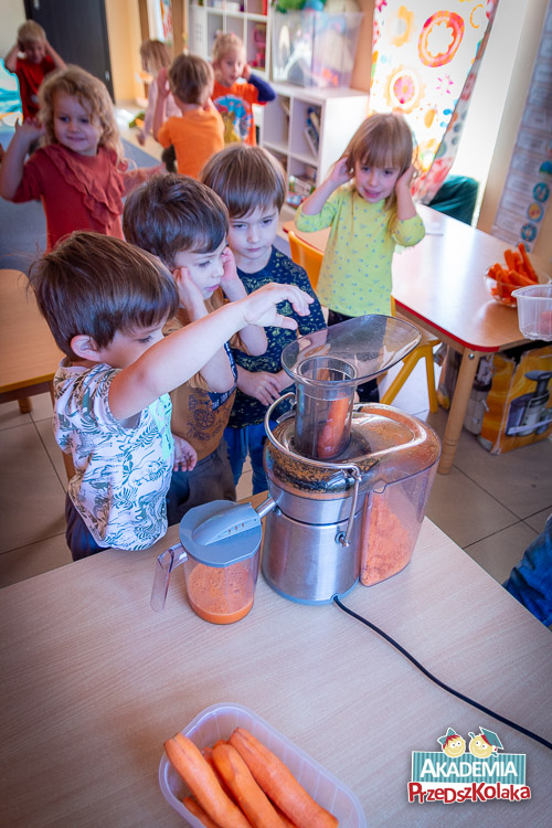 Przedszkolaki trzymają się za uszy. Uśmiechają się. Jeden z przedszkolaków wkłada marchewkę do działającej sokowirówki. Przedszkolaki pod okiem nauczycielek robią sok marchewkowo-owocowy.