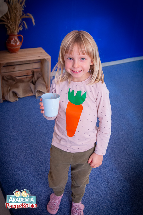 Przedszkolanka pozuje do zdjęcia. Dziewczynka stoi z kubkiem z sokiem. Szeroko się uśmiecha. Na koszulce ma wielką marchewkę.