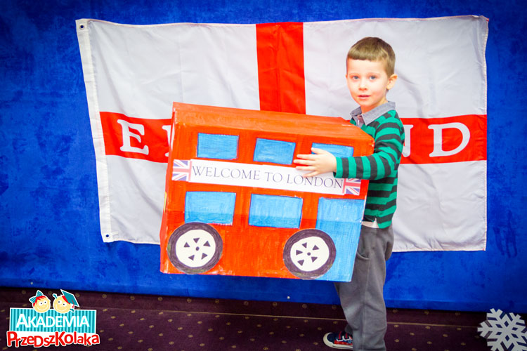 Przedszkolak na tle flagi angielskiej trzyma  makietę czerwonego, piętrowego  autobusu