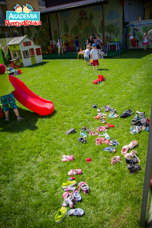 Rozrzucone na trawie placu zabaw buty dzieci. Dzieci bawią się boso.