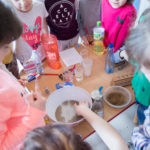 Dzieci stoją na około stołu. Do miski z wodą dodawane są różne substancje. Dzieci to obserwują.