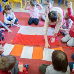 Przedszkolaki siedzą w kółku na dywanie. Po środku leży flaga Polski.