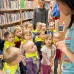 Grupa najmłodszych przedszkolaków stoi w bibliotece, wśród regałów z książkami. Pani bibliotekarka oprowadza i opowiada dzieciom o bibliotece.