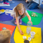 Przedszkolaki uczą się segregować śmieci. Na dywanie leżą rysunki śmietników. Przedszkolak odkłada plastikową butelkę na odpowiednie miejsce.