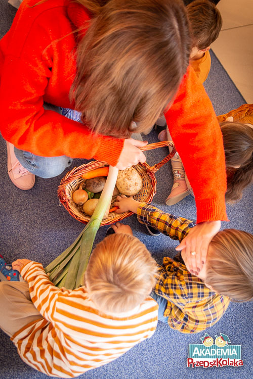 Przedszkolaki próbują wymacać marchewkę z kosza pełnego różnych warzyw. Nauczycielka zasłania chłopcu oczy ręką.