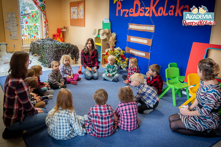 Przedszkolaki z nauczycielkami siedzą na dywanie. Na ścianie wisi napis Przedszkolna Akademia Rolnicza.