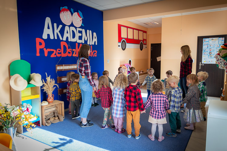 Sala przedszkolna w przedszkolu na Białołęce. Dzieci stoją w kółku, razem z nimi nauczycielki. Wszyscy trzymają się za ręce.