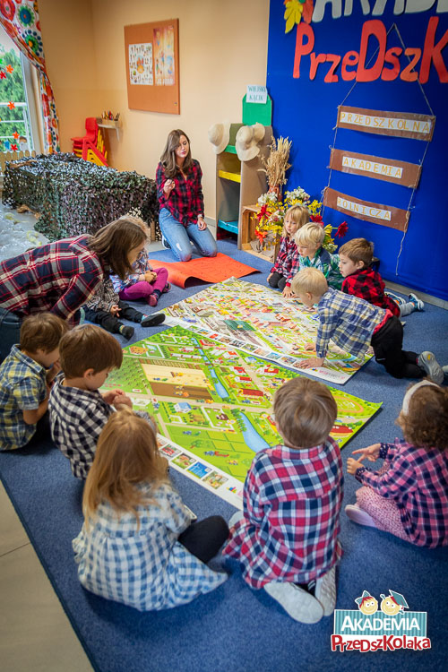 Przedszkolaki z przedszkola na Białołęce siedzą na dywanie, na około dwóch plansz z wizualizacjami wsi i miasta. Dzieci dokładnie oglądają, szczegółowe plansze.