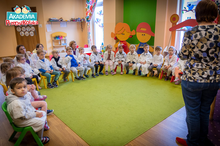Widok przedszkolaków siedzących w kółeczku. Wszystkie dzieci ubrane są w białe fartuchy. Prawdziwi mali naukowcy.