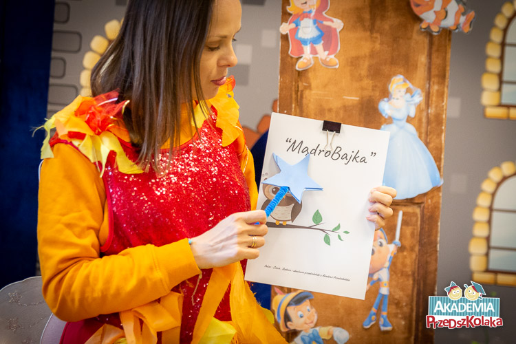Nauczycielka pokazuje magiczną, niebieską pałeczką okładkę MądroBajki której początek wymyśliły przedszkolaki. Dalszą część dzieci napiszą z rodzicami. Na okładce jest tytuł oraz rysunkowa sowa na gałązce. 
