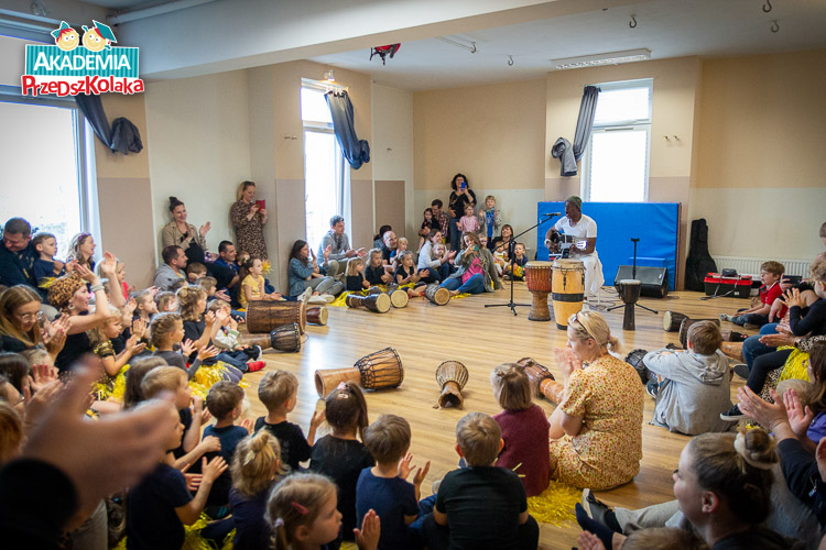 Ponownie sala przedszkolna na Białołęce. Widok ogólny całego wydarzenia. Dzieci z rodzicami klaszczą w dłonie. Gwiazda warsztatów gra i śpiewa.
