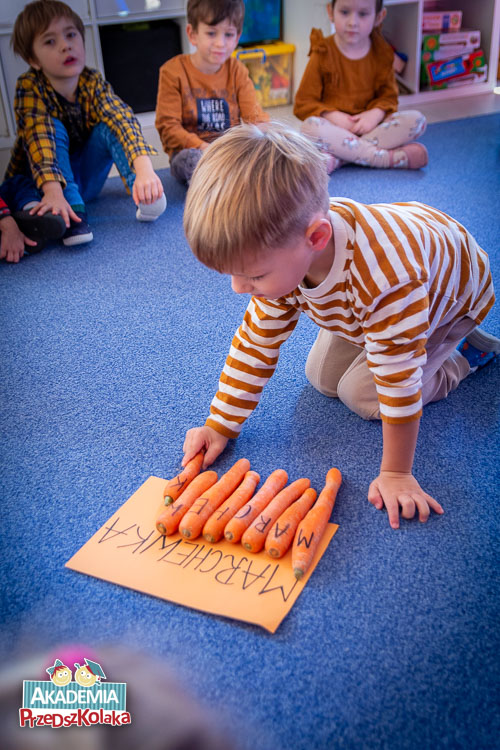 Przedszkolaki siedzą na dywanie. Jedno z dzieci układa marchewki z napisanymi literami w słowo MARCHEWKA. Warzywa układa na kartonie z napisem MARCHEWKA. 