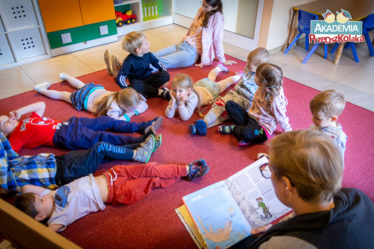 Nauczycielka czyta książkę, która znajdowała się na wcześniejszym zdjęciu. Dzieci leżą w różnych pozycjach na dywanie. Wsłuchane są w to co czyta nauczycielka o ewolucji.