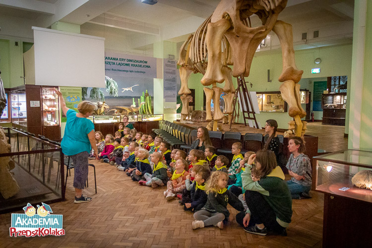 Przedszkolaki siedzą na ziemi z drewnianej klepki. Za plecami mają olbrzymią, 1:1 makietę szkieletu dinozaura. Pani przewodnik stoi przed siedzącymi przedszkolakami. 