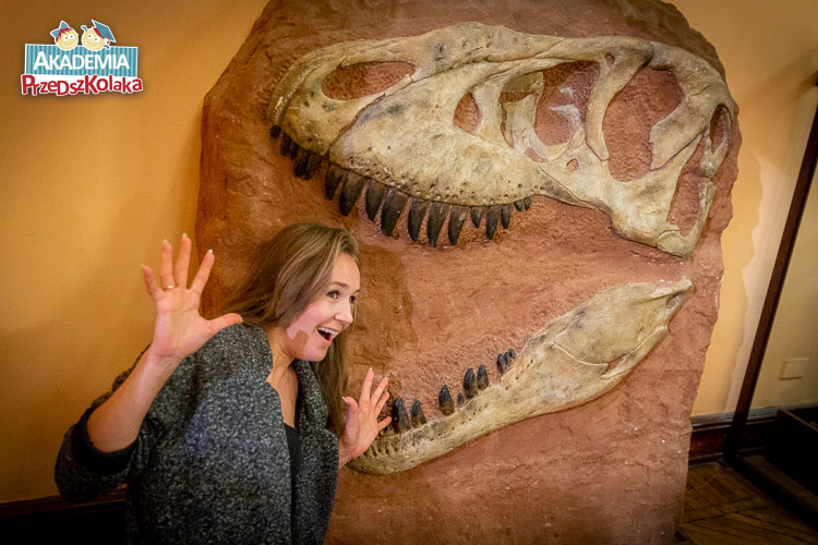 Skamielina w której zatopiony jest łeb jakiegoś drapieżnika. Nauczycielka udaje przerażoną, wykonując gest wkładania swojej głowy w rozwartą szczękę skamieliny dinozaura.