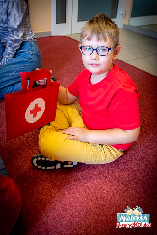 Portret przedszkolaka, który siedzi na dywanie i pokazuje do kamery swoją teczkę pracy. Teczka przypomina apteczkę. Jest czerwona.