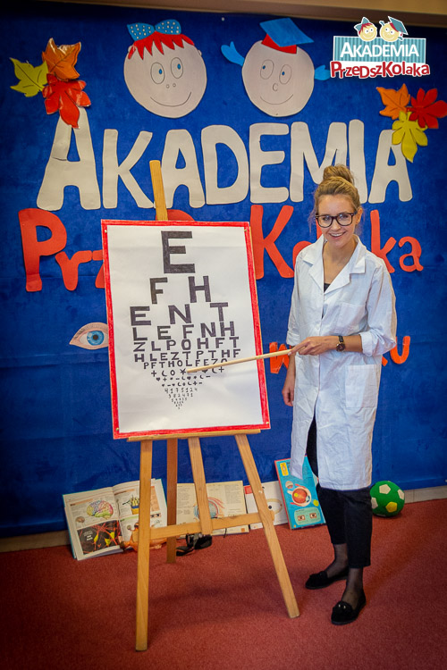Portret nauczycielki przebranej za lekarkę obok tablicy z różnej wielkości literkami.