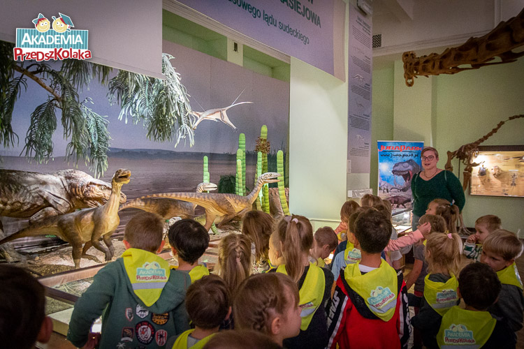 Przedszkolaki stoją przed makietą świata dinozaurów. Sztuczne modele bardzo realistycznie odwzorowują dinozaury. Widać rośliny, drzewa oraz latające zwierzęta, powieszone pod sufit.