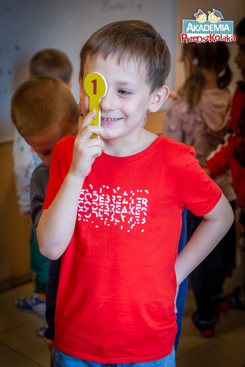 Portret przedszkolaka przykładającego do oka żółtego znaczka z czerwoną cyferką jeden. Chłopiec w czerwonym t-shert.