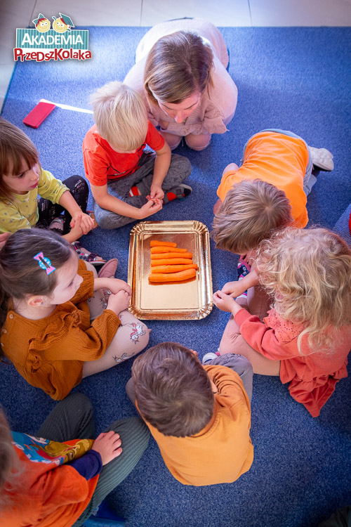 Dzieci na niebieskim dywanie siedzą na około złotej tacy z marchewkami. Warzywa ułożone są według długości kolejno.